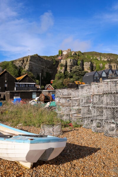 small white rowing boat and a stack of fishing nets on a stoney beach with some buildings behind and a large grassy cliff with a furnicular on it
