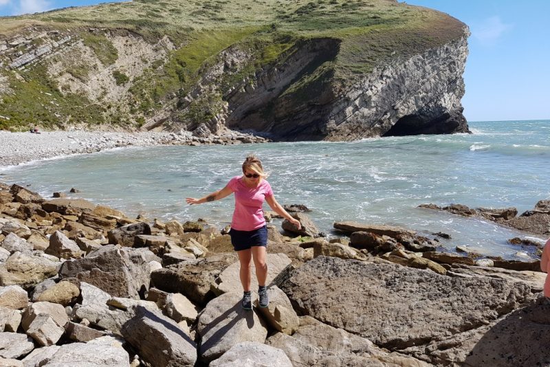 Emily walking on a rocky beach wearing shorts and a pink top 