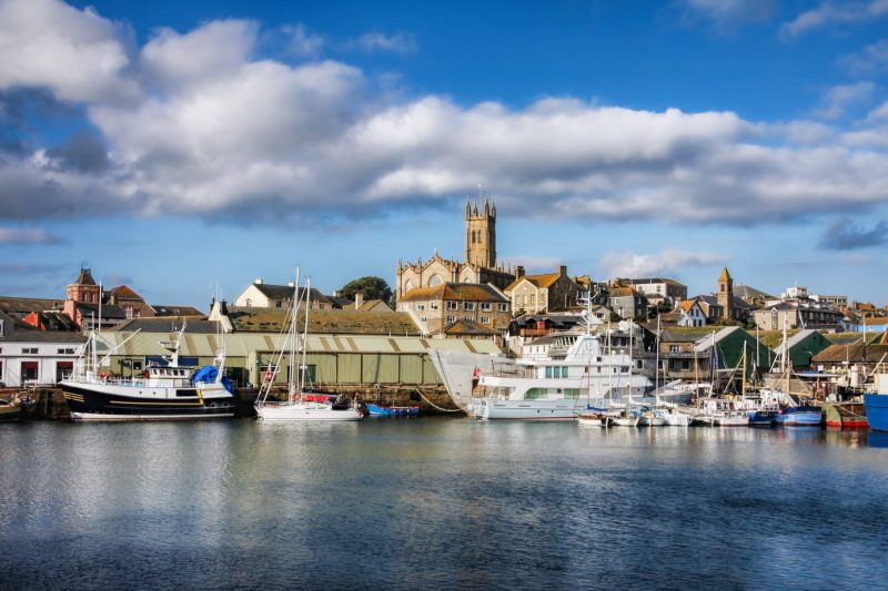 Penzance in Cornwall viewed from the water, with a harbour filled with small boats and the town on a hill behind, the church tower is on top of the hill against a blue sky filled with fluffy white clouds. 