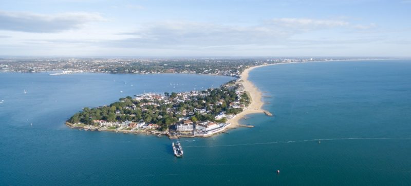 Aerial view of a densely populated coastal peninsula with buildings, a sandy beach along the right side, and a ferry near the shore. The background shows more coastline, buildings, and the horizon.