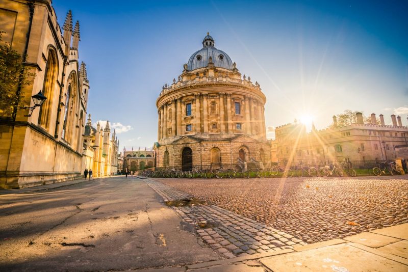 Radcliffe Science Library in Oxford with sunset flare - a circular building of beige stone with pillars around the outside and a grey slate domed roof. The sun is setting and the sky above is clear and blue. There is an empty cobbled street in front. Best Places to Visit in the South of England.