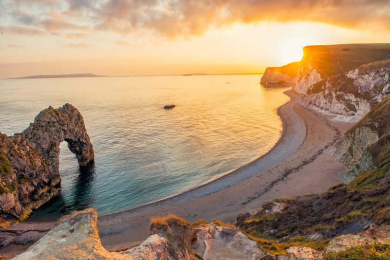 Durdle door viewed from the cliff, a grey rock arch over the sea beside a large bay with an empty shingle beach and distant white cliffs with the sun setting behind them and orange sky overhead. On the south coast of England,