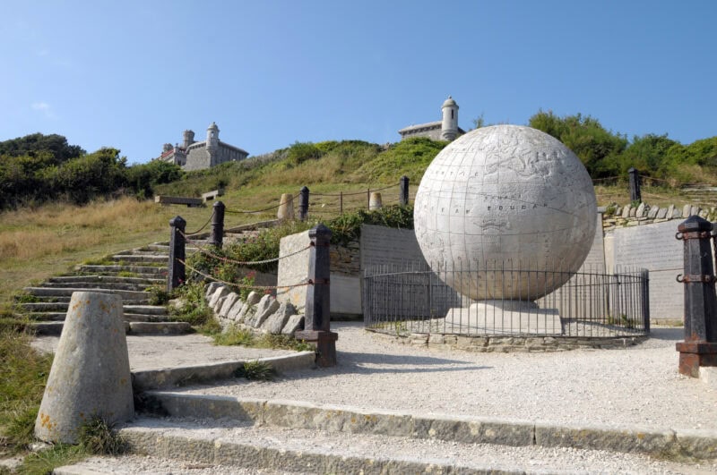 Large stone globe made from light grey stone on a pedastal at the foot of a grassy hill