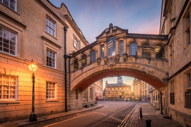 Bridge of sighs with the Sheldonian theatre background and street lamp foreground during twilight in Oxford. stone bridge running between two buildings on the second floor above a road.