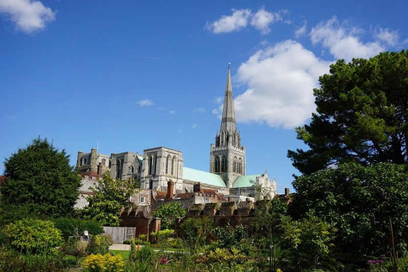 Grey stone church tower in Chichester with green leafy trees in front and clear blue sky above.