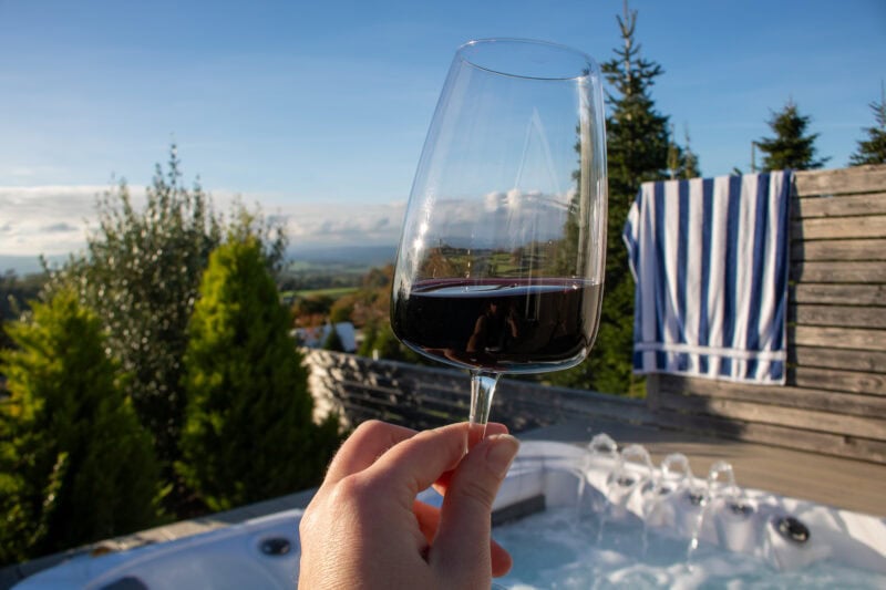 Hand holding a glass of red wine in front of an outdoor hot tub