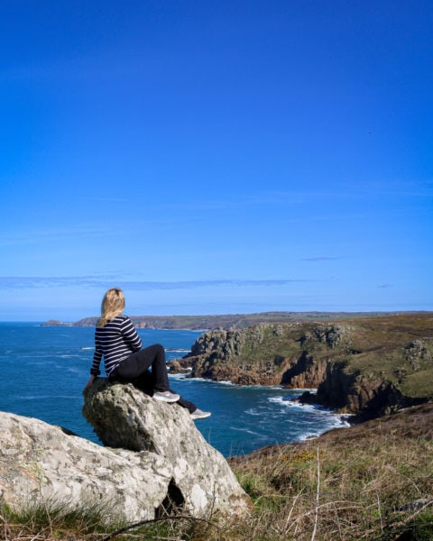 emily wearing black trousers and a navy and white striped long sleave top is sitting on a grey rock on the Penwith Coast in Cornwall looking out at rocky grassy cliffs and bright blue sea on a very sunny day with blue sky overhead.