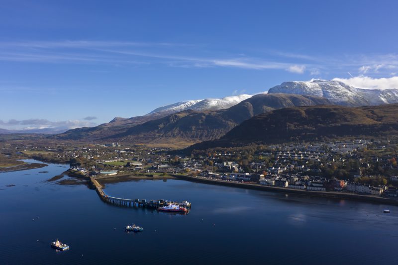 aerial view of Fort William town in Scotland next to a large blue lake with a snowcapped mountain behind on a sunny day with clear blue sky above