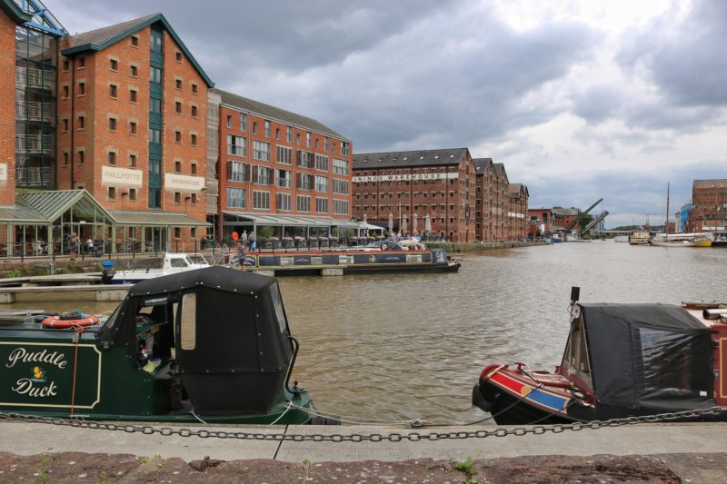 the edges of two canal boats against a walkway with a brown river behind them lined with large red brick warehouses - gloucester docks