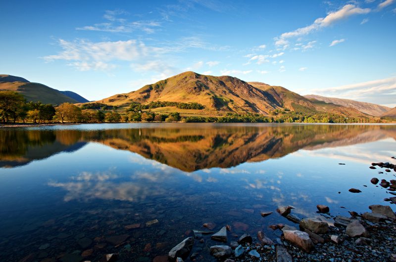 very calm flat lake with a hill on the far side reflected in the water on a sunny day with blue sky above - Buttermere Lake District England UK