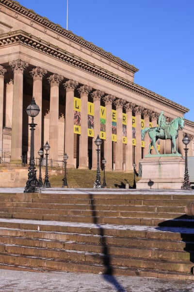 Exterior of a grand neoclassical building built from beige coloured stone with many columns. There are yellow squares with black letters in between the columnns which read: Liverpool Music City.