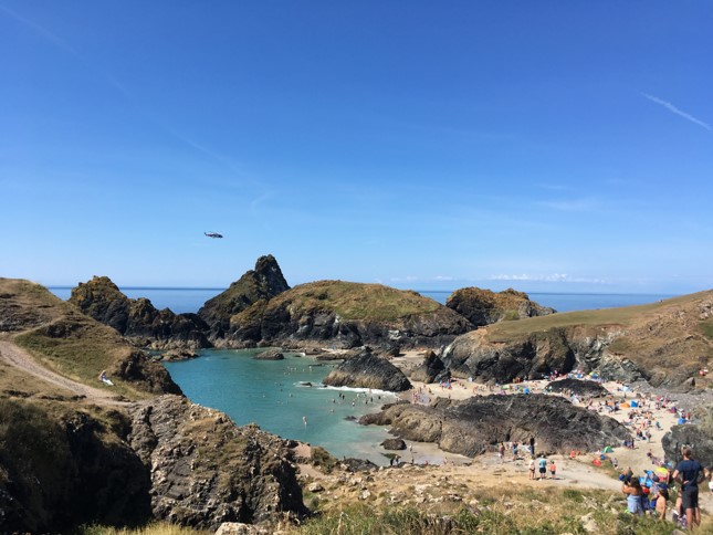 View of Kynance Cove on the Lizard Peninsula in Cornwall England - there is a bay with bright turquoise water and a sandy beach surrounded by jagged grey rocks on a very sunny day with clear blue sky above