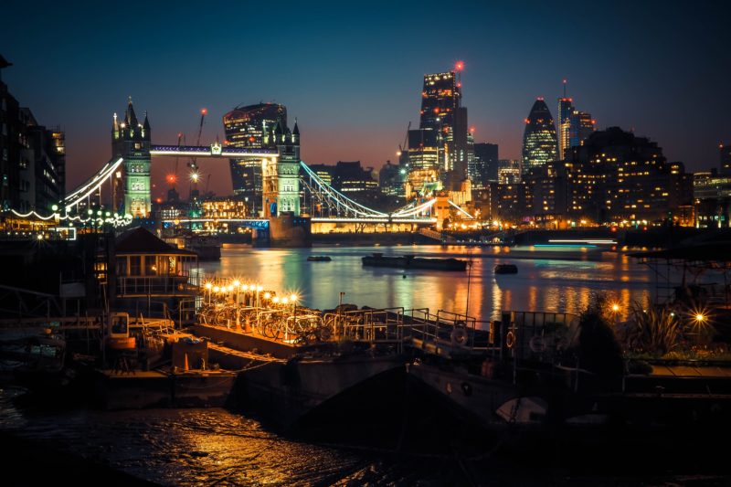 London Tower Bridge at Night viewed from the south bank. the bridge has a stone tower on either side, lit up with yellow and blue lights against the night sky with the skyscrapers of london in the background and all the lights reflected in the river thames. best cities in uk for nightlife.