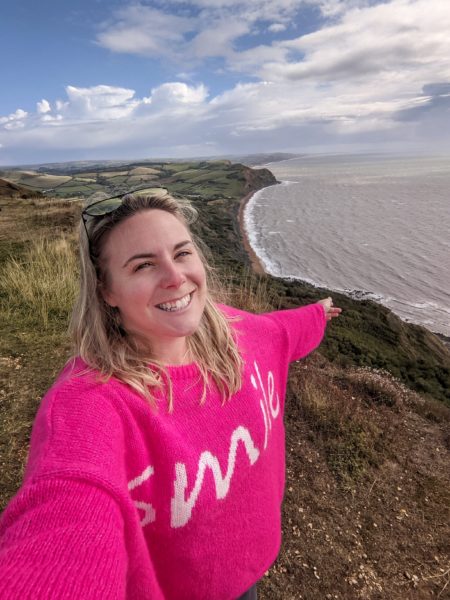 selfie of emily wearing a pink jumper that says Smile, standing on top of a cliff