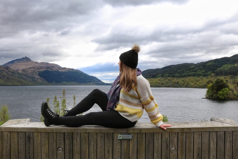 emily wearing a yellow wool jumper and black wool hat, sat on a wall next to a loch in Scotland | Solo Female Travel in the UK