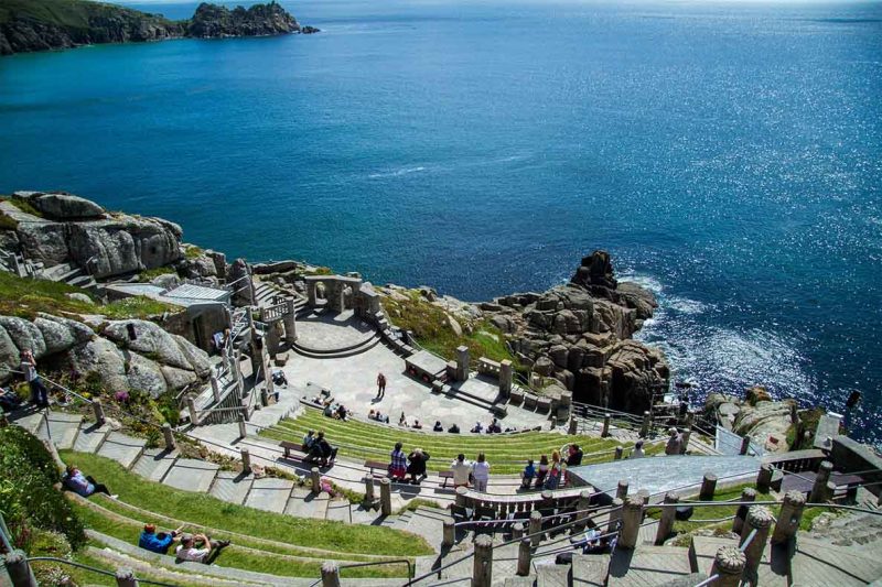 Open-air theater built into cliffs overlooking the sea. Stone seating in a semi-circle with grass and stone steps, leading down to the stage. The backdrop includes the expansive blue sea, rocky cliffs, and a clear sky. People are seated, enjoying a unique and scenic location.