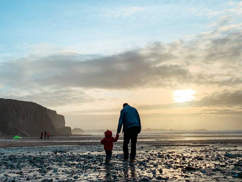 Man and toddler holding hands and walking on a beach just before sunset with cliffs silhouetted in the distance, taken in Newquay Cornwall in the South West of England