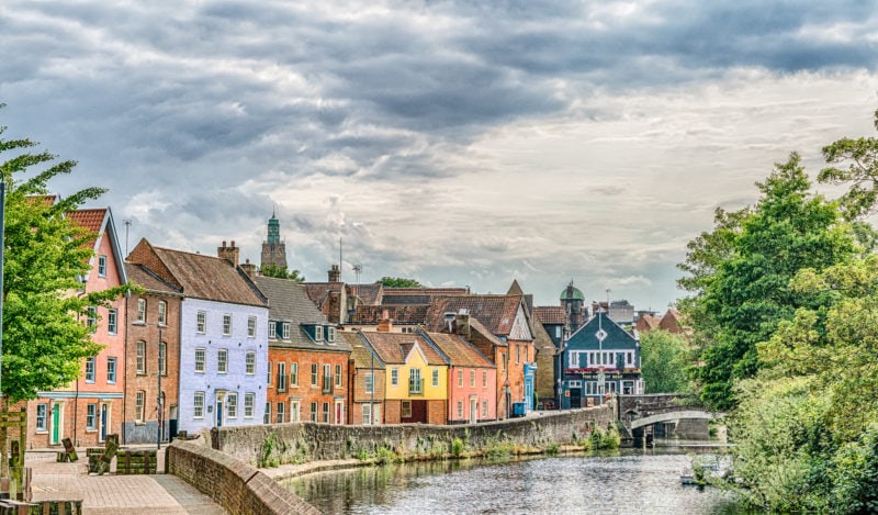 looking down a calm, grey river in Norwich with a row of stone buildings with grey tiled roofs each one painted a different colour, the scene is framed by green leafy trees on either side and its a grey cloudy day