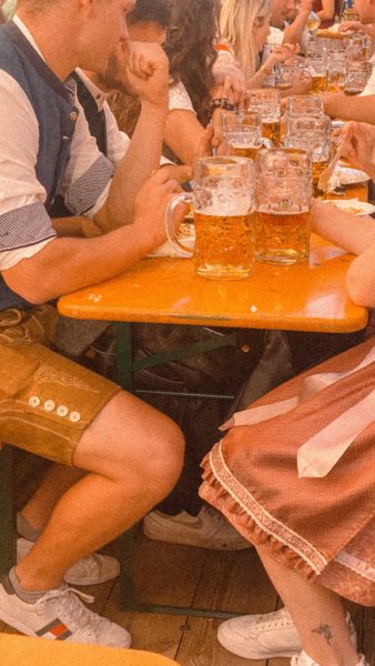 a groupd of people sitting at a long wooden table wearing traditional lederhosen and dirndl and drinking beer. the image is cropped so that you can't see their faces. Oktoberfest events in the UK.