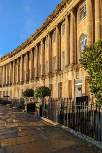 exterior of a three storey georgian townhouse in Bath Englamnd built from beige coloured stone with stone collonades on the upper two floors. there are two small green bushes outside the entrance gate.