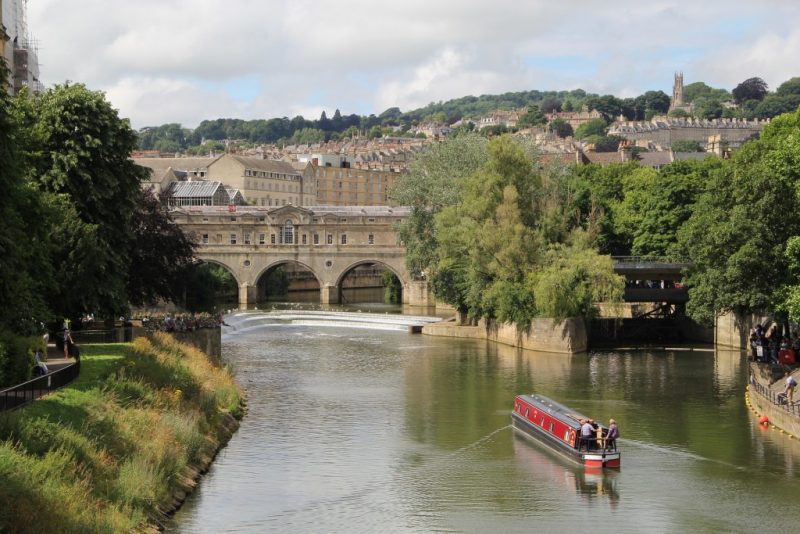 Avon River in Bath with a red wooden narrow boat sailing up the river towards a covered bridge built from beige stone. There are green leafy trees on both banks and Bath city ckyline on the hill behind.